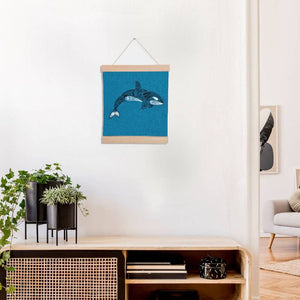 A wooden banner holding a teal square of fabric, set against a white background. The fabric is embroidered with a paisley style orca design.