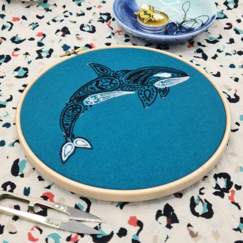 An embroidery hoop is lying on a pattern piece of fabric. The hoop is holding a teal piece of fabric, embroidered with a paisley style Killer Whale design.
