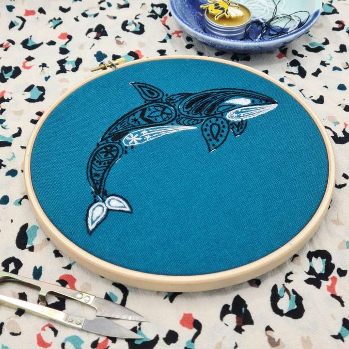 An angled photo of a black and white Orca embroidered on a teal fabric held in a wooden hoop