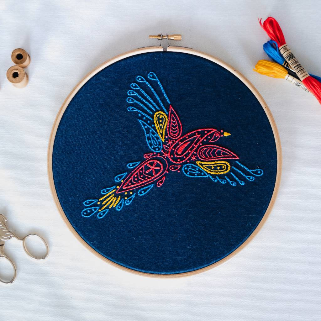 A finished parrot embroidery design on navy fabric, with thread and accessories surrounding it. Made using this parrot embroidery kit product.