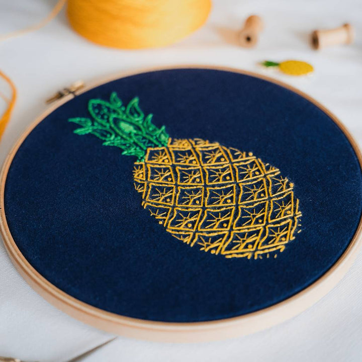 An Angled photo of a finished Pineapple Embroidery design on navy fabric, with yellow thread and a needleminder in the background. Made using this Pineapple Embroidery Kit product.