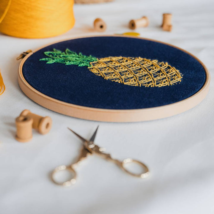 An Angled photo of a finished Pineapple Embroidery design on navy fabric, with scissors in the foreground and yellow thread in the background. Made using this Pineapple Embroidery Kit product.