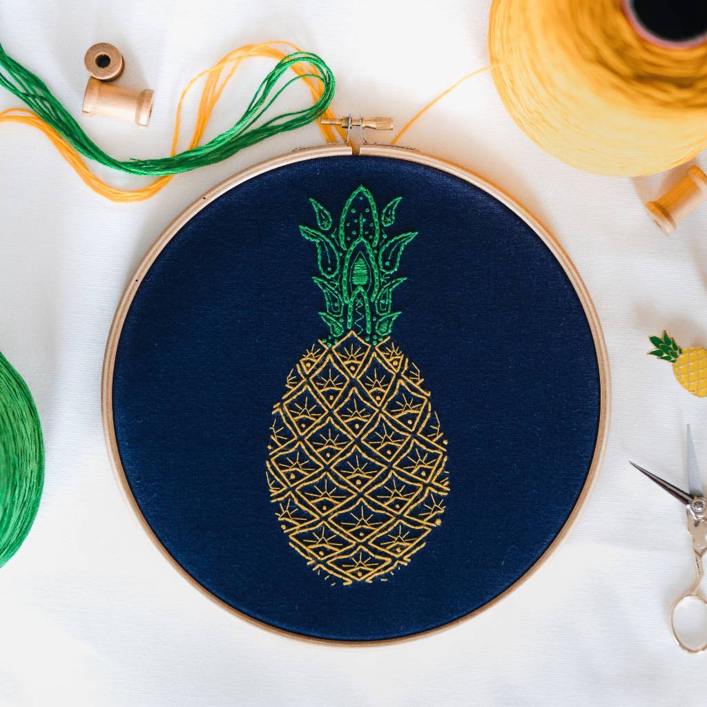 A finished Pineapple embroidery design on navy fabric, with decorative accessories and yellow and green thread surrounding it. Made using this Pineapple Embroidery Kit product. 