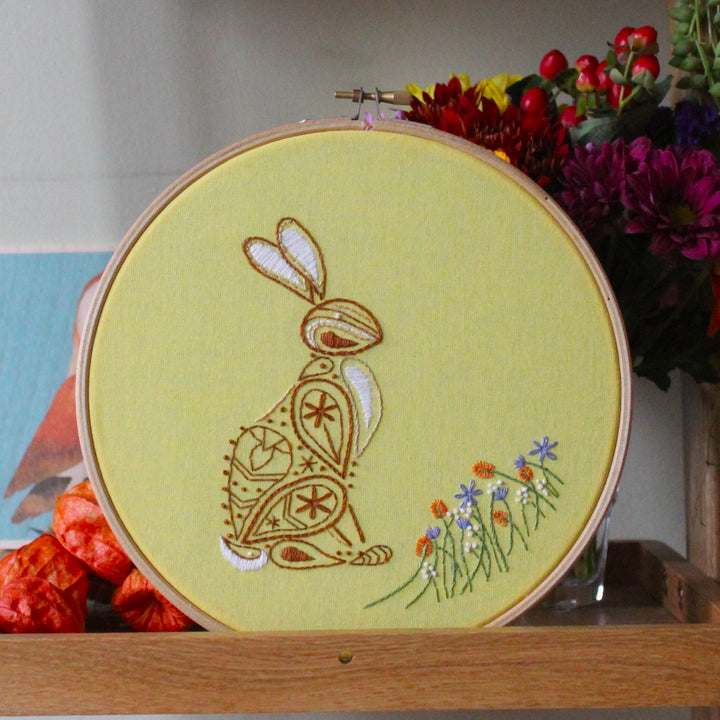 An embroidery hoop holding light yellow fabric standing on a wooden shelf. The fabric is embroidered with a paisley style rabbit and a row of flowers.