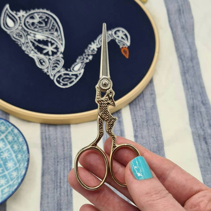 A hand holding up a pair of rabbit embroidery scissors with a swan embroidery hoop in the background.