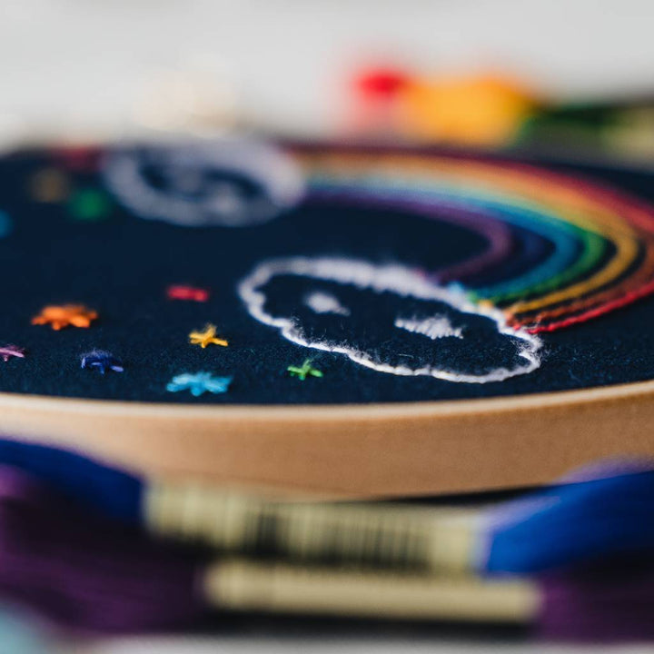 An angled close up photo of a finished Rainbow embroidery design on navy fabric with thread in the foreground. Made using this rainbow embroidery kit product.