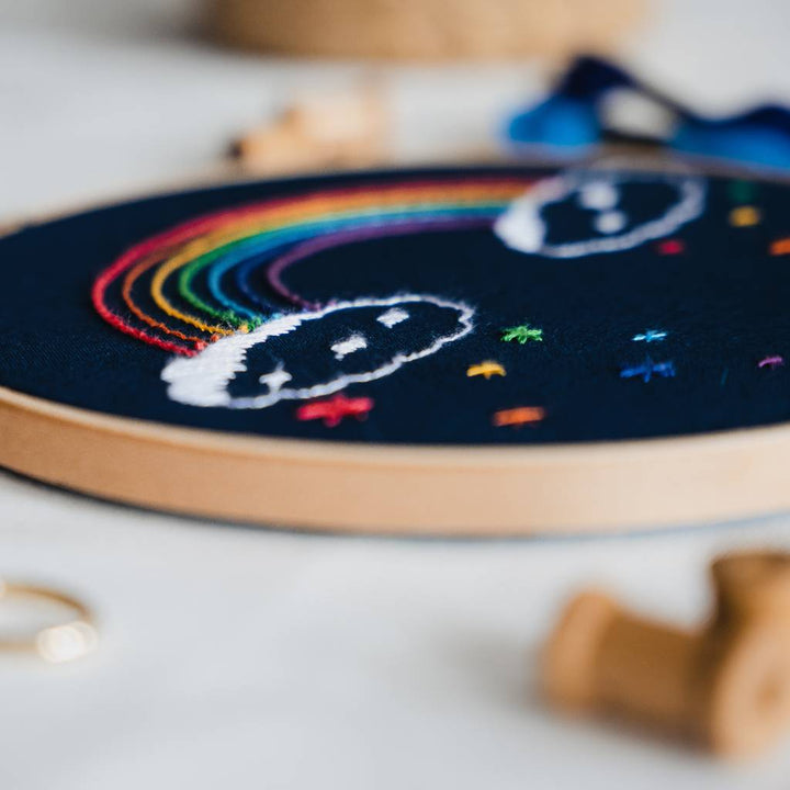 An angled photo of a finished Rainbow embroidery design on navy fabric with thread spools in the foreground. Made using this rainbow embroidery kit product.