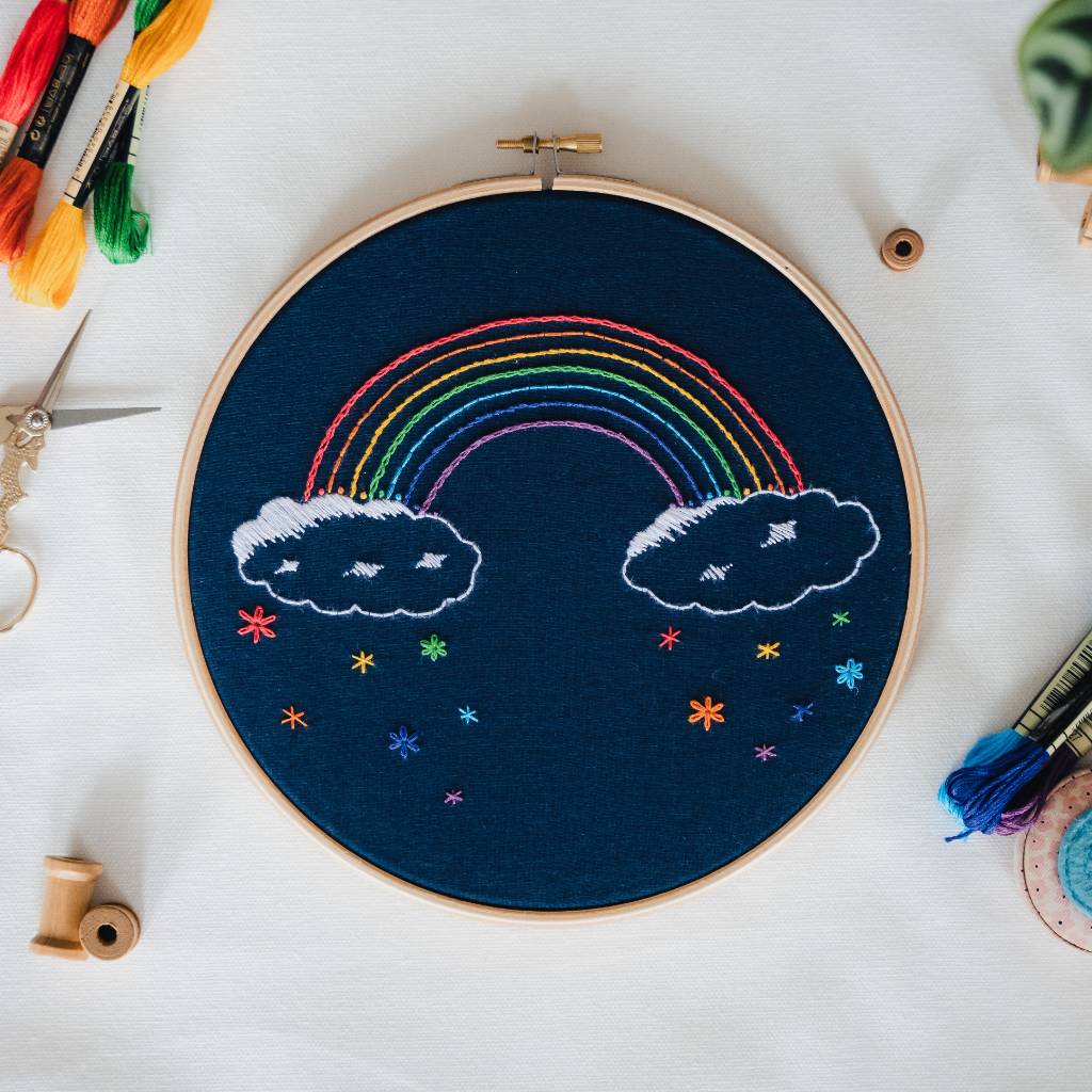 A finished rainbow embroidery design on navy fabric, with thread and accessories surrounding it. Made using this Rainbow embroidery kit product.