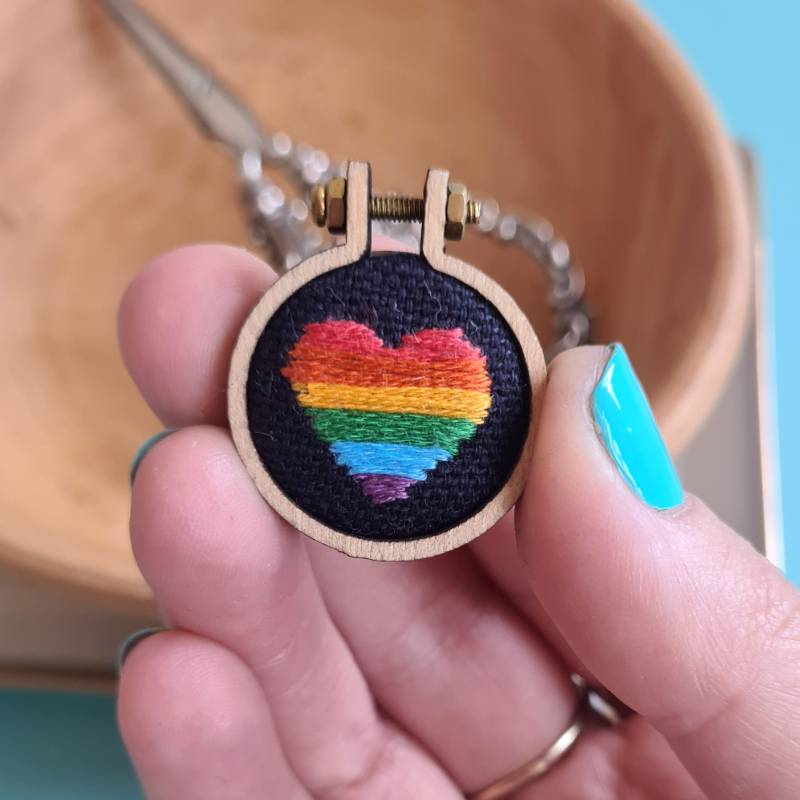 A rainbow heart stitched into fabric in a charm held by fingers
