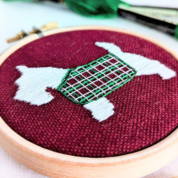 Very close up front left angled photo of a white scottie dog with a tartan jacket embroidered on red fabric