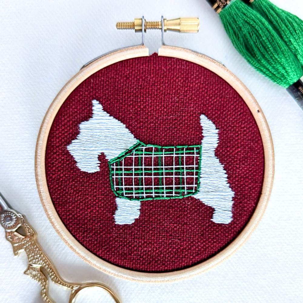 Close up photo of a white scottie dog with a tartan jacket embroidered on red fabric 