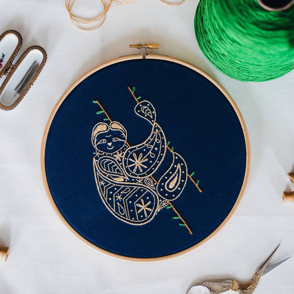 A finished sloth embroidery design on navy fabric, with accessories and thread surrounding it. Made using this sloth embroidery kit product.