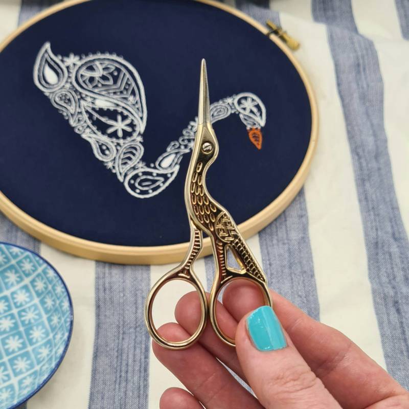 A hand holding up a pair of stork embroidery scissors, with a swan embroidery hoop in the background.