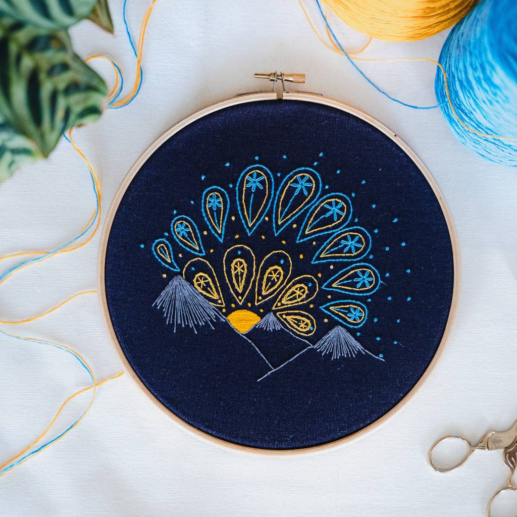 A finished sunrise embroidery design on navy fabric, with thread and leaves above it and scissors below it. Made using this sunrise embroidery kit product.