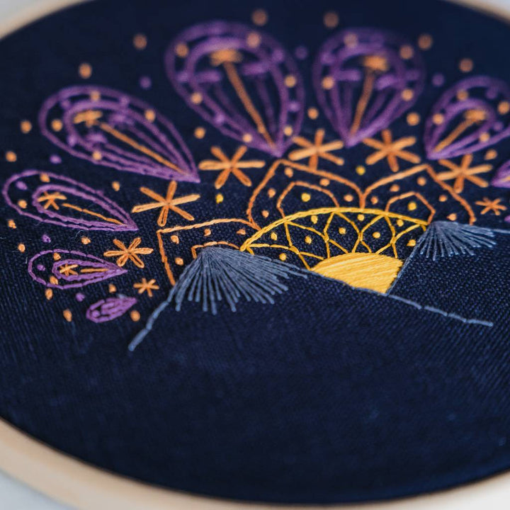 An angled photo of a sunset embroidery design with the sun at the bottom, on navy fabric. Made using this sunset embroidery kit product.