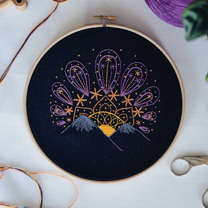 A finished sunset embroidery design on organic navy fabric, with thread and accessories surrounding it. Made using this sunset embroidery kit product.