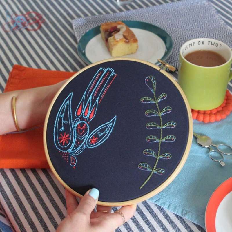 A blue and red may morris inspired embroidery design flying next to an embroidered branch