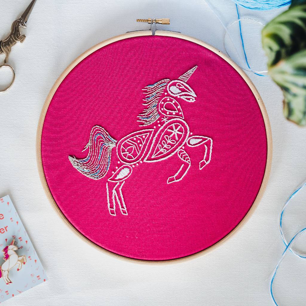 A finished unicorn embroidery design on pink fabric with unicorn scissors and needleminder on the left and thread and leaves on the right. Made using this unicorn embroidery kit product.