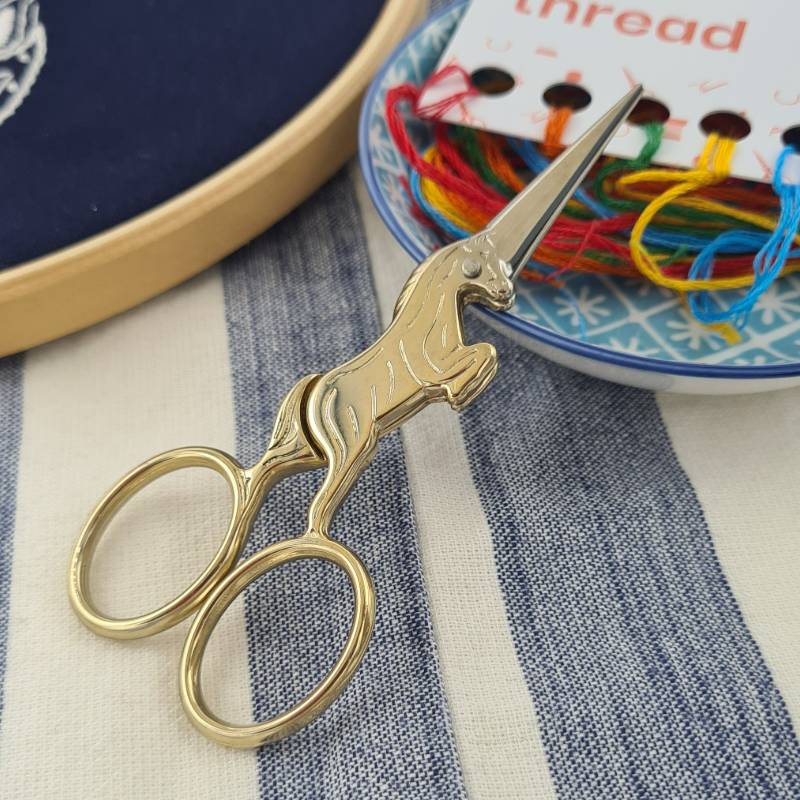 A pair of gold unicorn embroidery scissors resting on a small bowl containing colourful embroidery thread.