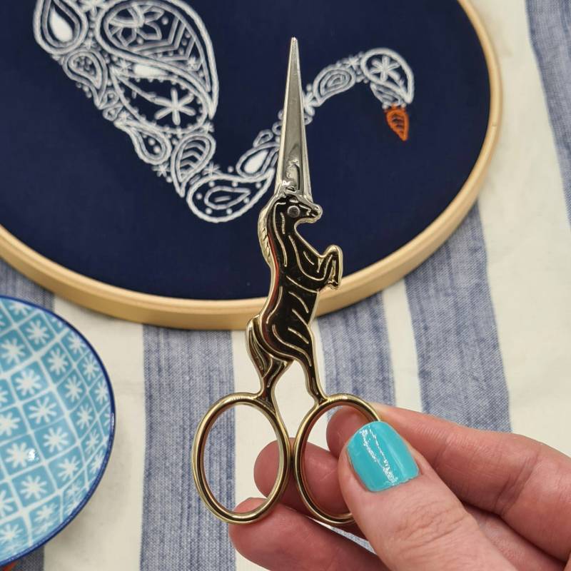 A hand holding up a pair of gold unicorn embroidery scissors, with a swan embroidery hoop in the background.