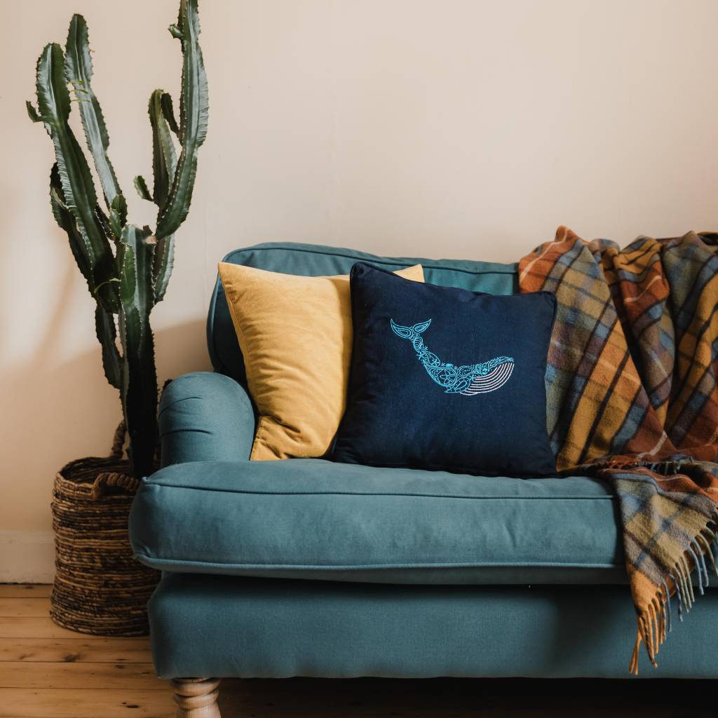 A finished whale embroidery design on an organic navy cushion, on a sofa with a cactus behind it, made using a whale cushion embroidery kit product.