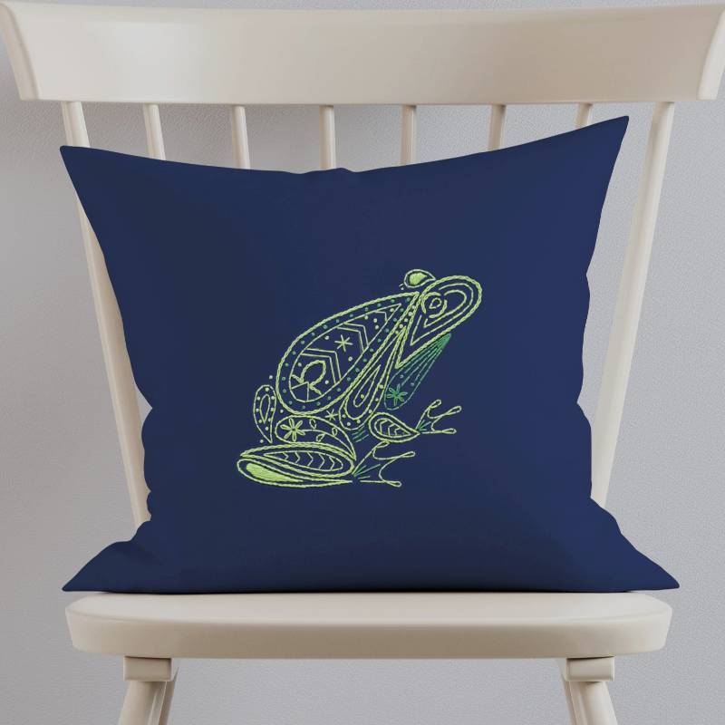 A navy blue cushion embroidered with a green frog design sits on a white wooden chair.