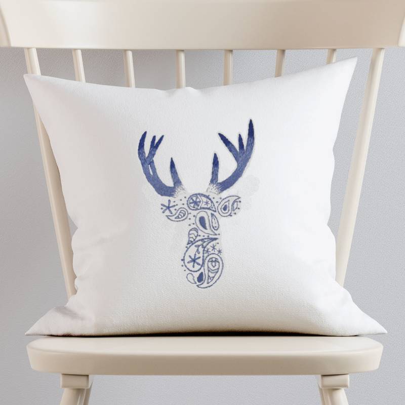 A white cushion sitting on a white wooden chair. The cushion is hand embroidered with a paisley deer pattern in blue and purple thread.