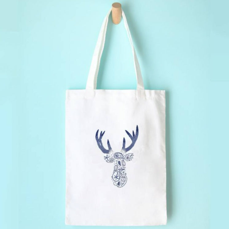 A white tote bag hanging on a wooden peg against a blue background. On the tote bag, a blue paisley deer design is embroidered.