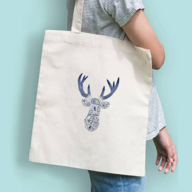 A person standing side on, carrying a cream tote bag on their shoulder. The tote bag displays an embroidered stag design.