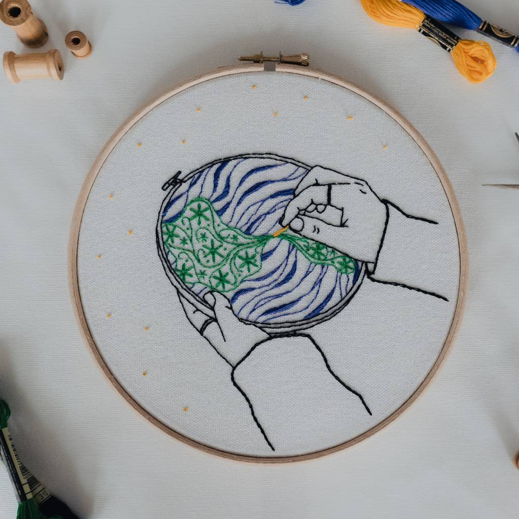 an embroidery hoop on a cream background stitched with a design showing hands holding a globe