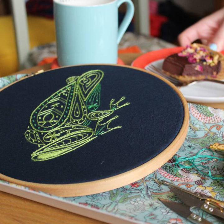 An embroidery hoop holding navy blue fabric stitched with a green frog design rests on a blue patterned tray, with a plate of cake and mug in the background.