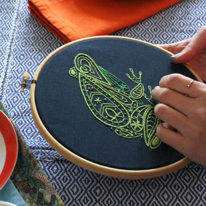 Hands holding an embroidery hoop holding navy blue fabric stitched with a green frog design, against a blue and white pattern tablecloth.