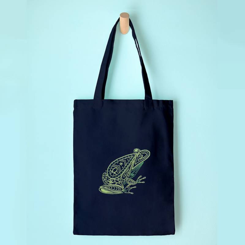 A navy blue tote bag hangs on a wooden peg against a light blue background. The bag is embroidered with a green, paisley style frog design.