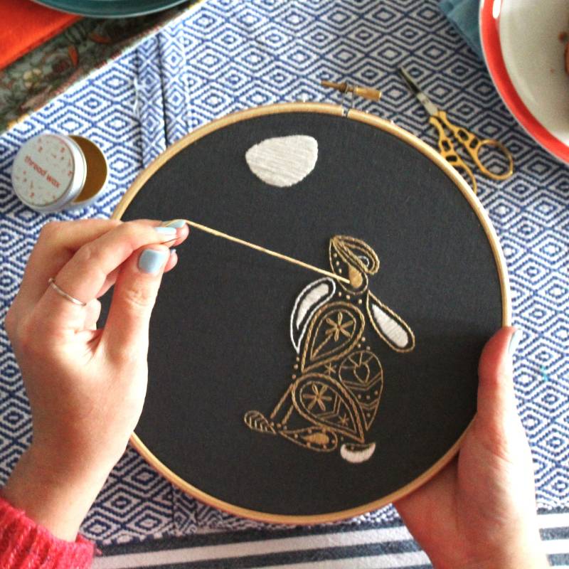 A pair of hands holding an embroidery hoop, stitching a paisley style hare design.
