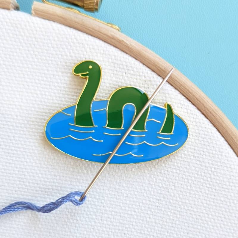 An embroidery needle rests on a Loch Ness monster magnet, against a white fabric background.