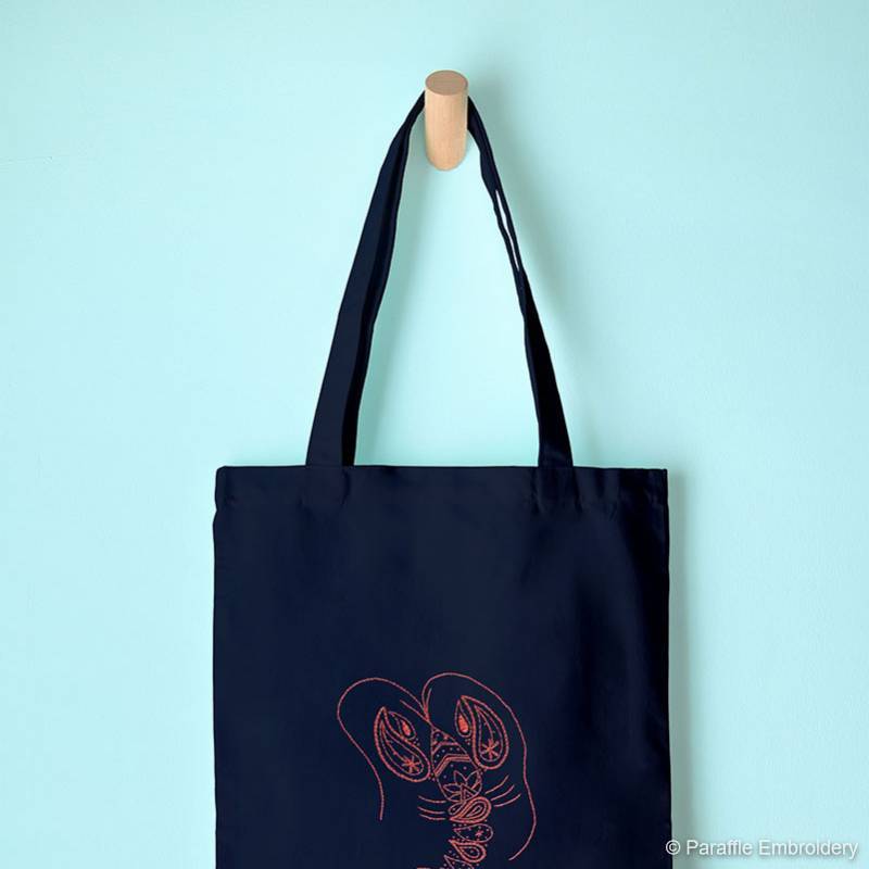 Lifestyle view  of paisley lobster hand embroidery on navy tote bag hung from hook 