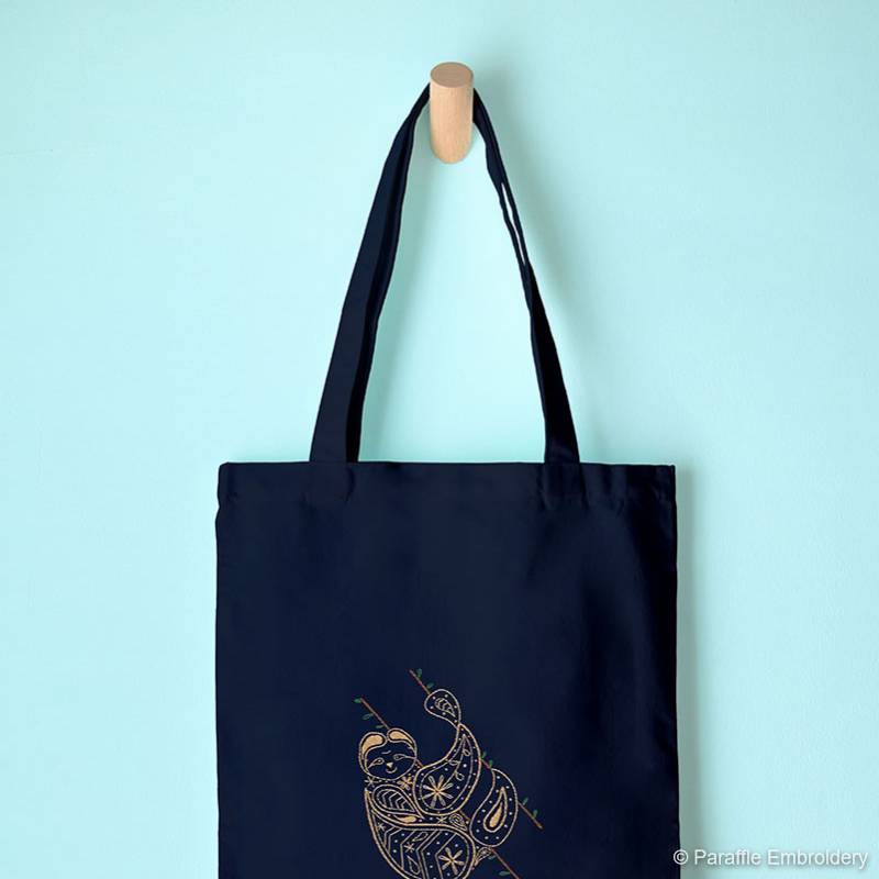 Lifestyle view of paisley sloth embroidery on navy tote bag hung from display hook