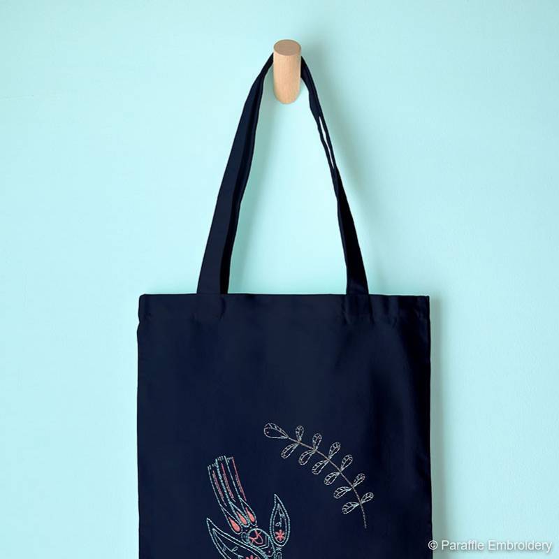 Lifestyle view of paisley swallow hand embroidery on navy tote bag hung from wooden hook