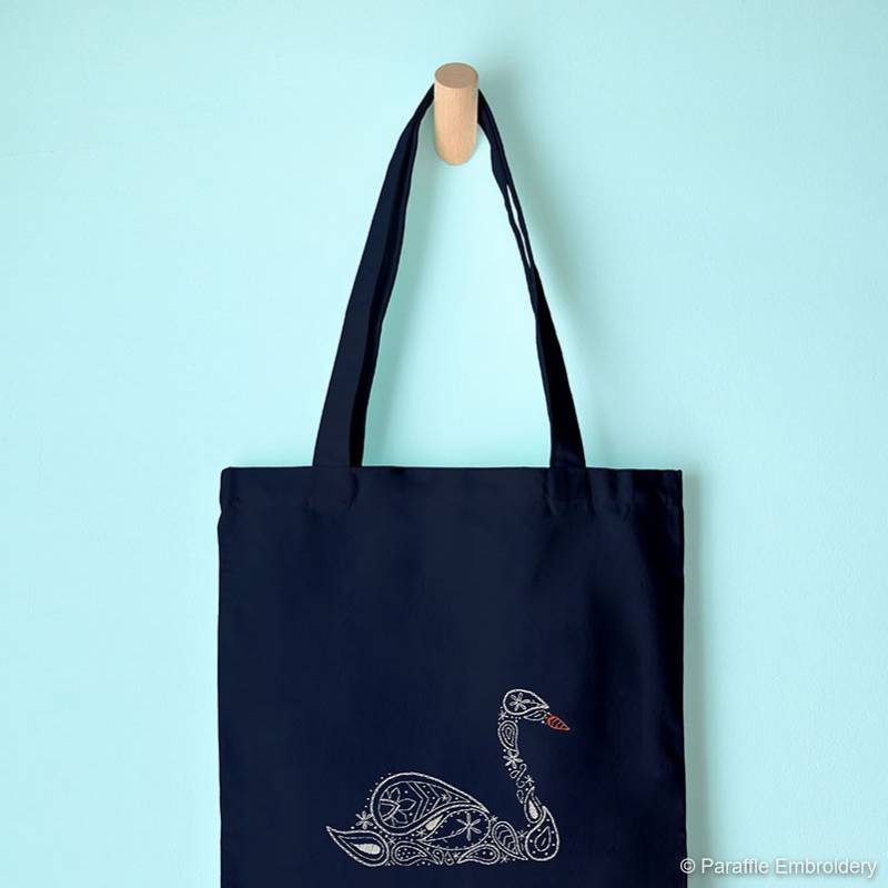 Lifestyle view of paisley swan embroidery on navy tote bag hung from display hook