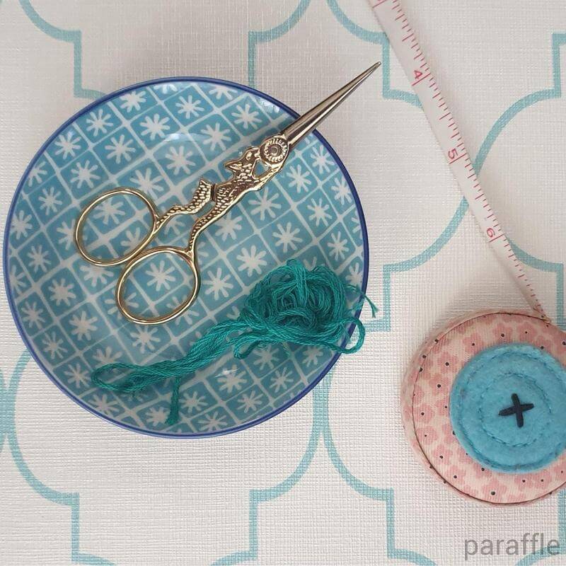 A pair of gold rabbit embroidery scissors resting in a small bowl containing teal thread, with a tape measure on the side.