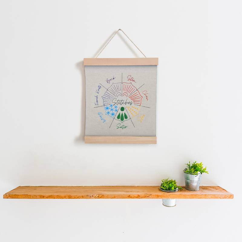 A piece of cream fabric stitched as an embroidery sampler hangs from a hook, above a shelf with two small plants.