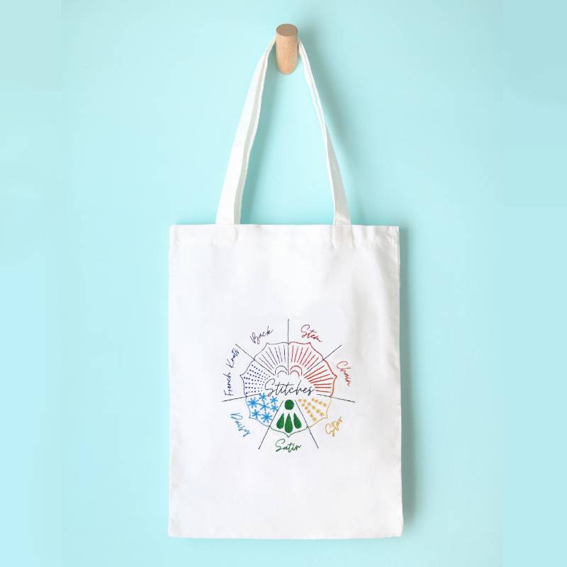 A cream tote bag hangs against a blue wall. The tote bag is embroidered with a sampler embroidery design in rainbow colours.
