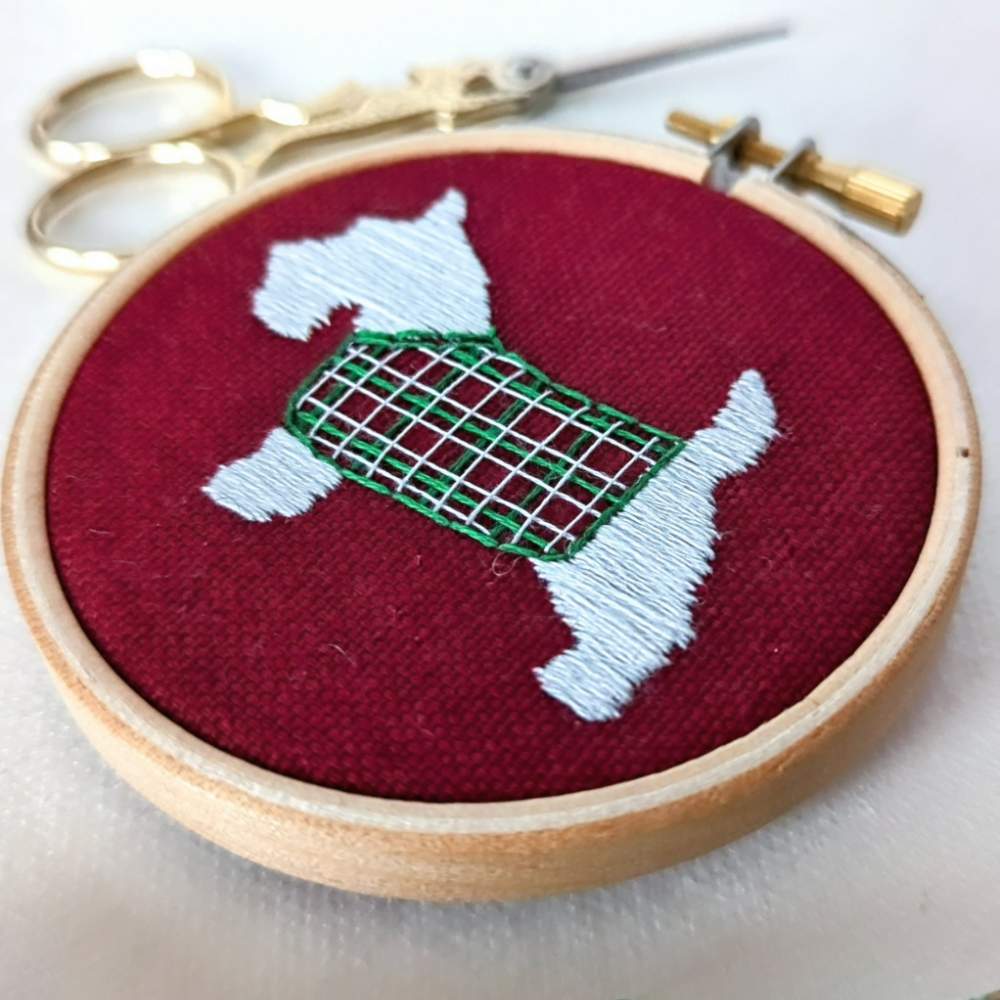 Close up front right angled photo of a white scottie dog with a tartan jacket embroidered on red fabric