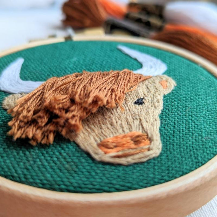 Close up of a Highland Cow stitched on green fabric with brown and beige embroidery thread.