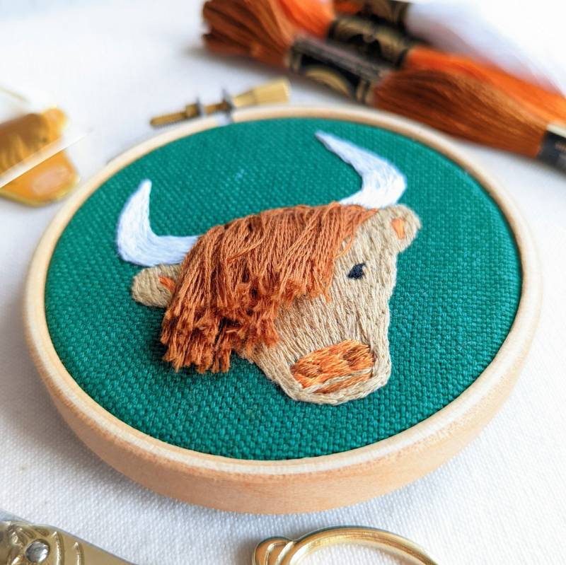 Side view of a small embroidery hoop, containing green fabric stitched with a highland coo design in brown embroidery thread.