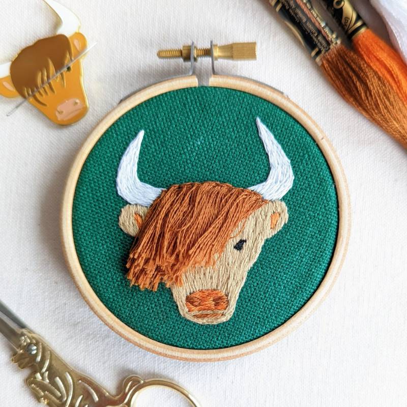 A bird&#39;s eye view of a small embroidery hoop containing green fabric, embroidered with a highland cow design.