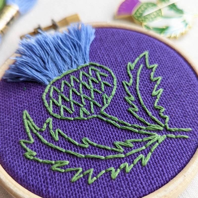 Close up of purple fabric embroidered with a lilac and purple Scottish thistle design.