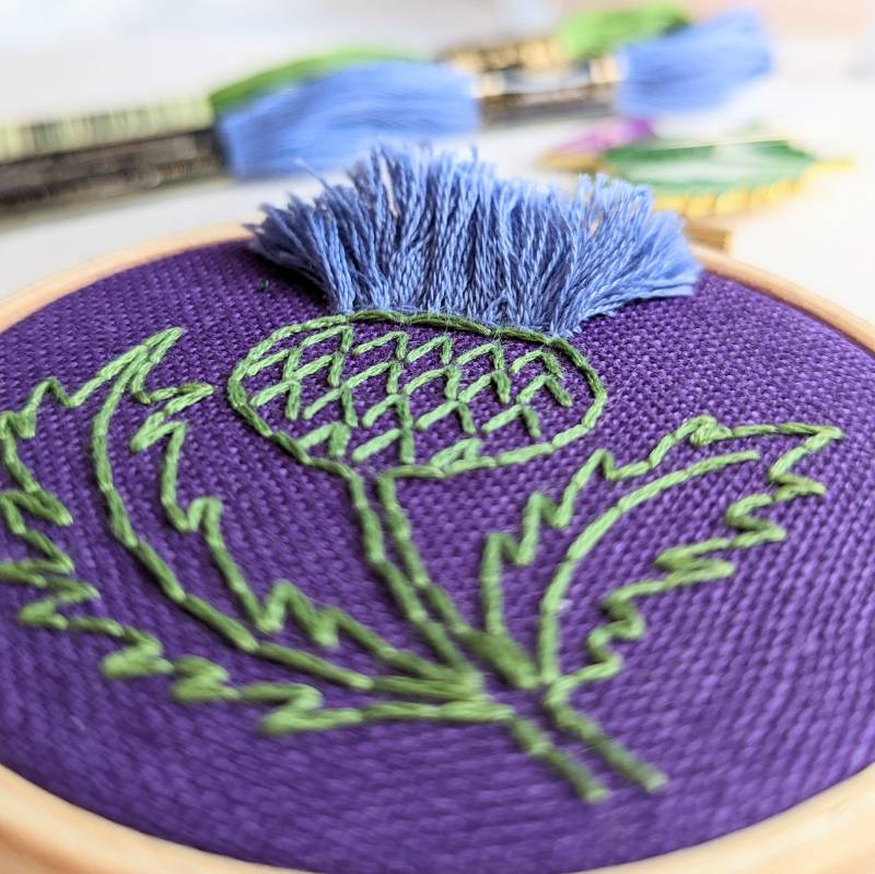 Side view of a small embroidery hoop containing purple fabric, stitched with a simple thistle design in lilac and green thread.