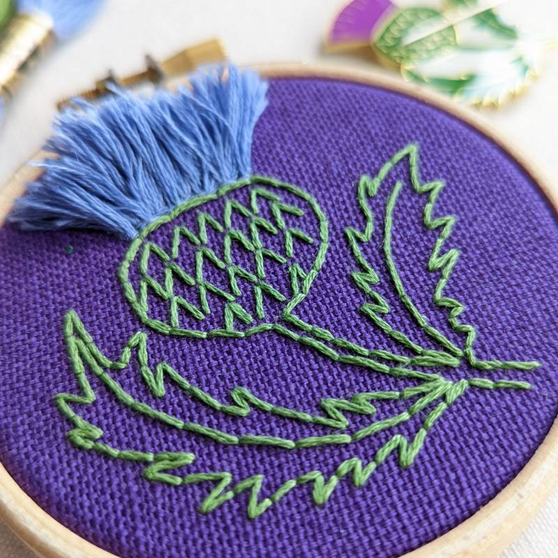 Close up of purple fabric embroidered with a lilac and purple Scottish thistle design.