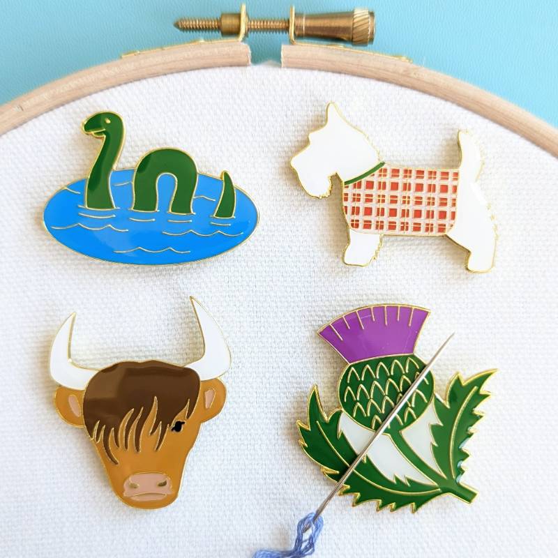 Four magnets - showing a Loch Ness monster, Highland terrier, Highland Coo, and Thistle, against a white fabric held in a wooden embroidery hoop.